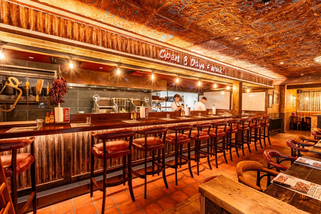A row of bar chairs at the Chefs Bar with two chefs working in the kitchen at the Swindon Hickorys Smokehouse.